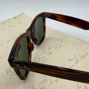 May include: Close-up of Ray-Ban sunglasses with a tortoiseshell frame and green lenses. The Ray-Ban logo is visible on the temple. The sunglasses are resting on a piece of paper with handwritten text.