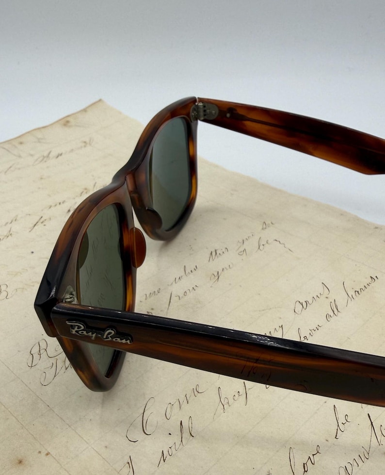 May include: Close-up of Ray-Ban sunglasses with a tortoiseshell frame and green lenses. The Ray-Ban logo is visible on the temple. The sunglasses are resting on a piece of paper with handwritten text.