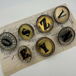Könnte beinhalten: Eine Sammlung von Vintage-Buchstabenanhängern mit schwarzen Buchstaben auf goldenem oder silbernem Hintergrund. Die Buchstaben sind in runden, klaren Glas- oder Kunststoffgehäusen eingeschlossen. Die Anhänger sind auf einem Vintage-Beleg angeordnet.