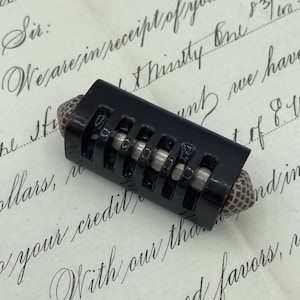 May include: A black, vintage bead with a rectangular, slotted design. The bead has a cylindrical shape with a patterned end. The background is a handwritten letter.