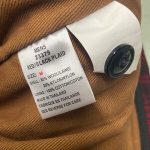 May include: Close-up of a tag from a red and black plaid jacket. The tag reads "MENS 23379 RED/BLACK PLAID SIZE: M SHELL: 80% WOOL/LAINE/ 20% NYLON/NYLON LINING: 100% COTTON/COTON MADE IN THAILAND". A black button is visible.
