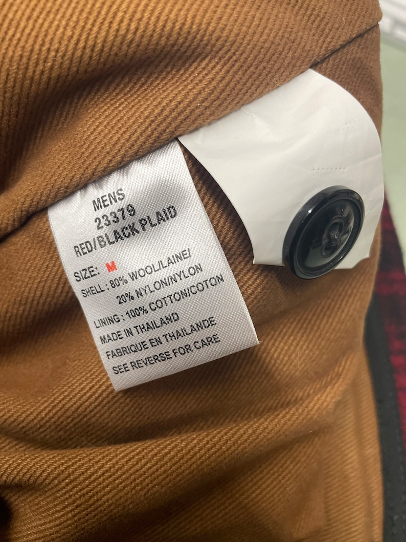 May include: Close-up of a tag from a red and black plaid jacket. The tag reads "MENS 23379 RED/BLACK PLAID SIZE: M SHELL: 80% WOOL/LAINE/ 20% NYLON/NYLON LINING: 100% COTTON/COTON MADE IN THAILAND". A black button is visible.