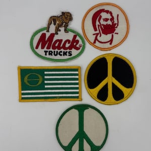 May include: A collection of five embroidered patches including an oval Mack Trucks patch with a bulldog, a circular patch with a red bearded figure, a rectangular green and white flag patch with a symbol, and two circular peace sign patches.