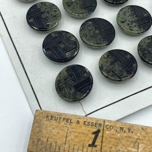 Puede incluir: Una colección de 16 botones negros y verdes con un patrón geométrico. Los botones están dispuestos en filas sobre una superficie blanca. Los botones tienen un diámetro de aproximadamente 2,5 cm.