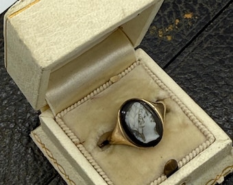 Op de afbeelding: Een gouden ring met een zwarte ovale steen met een wit camee portret, gepresenteerd in een open crèmekleurige sieradendoos met gouden randen. De ring ligt op een beige fluwelen kussen.