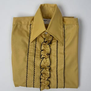 Pode incluir: Camisa mostarda com botões, gola pontuda e acabamento preto. A camisa apresenta uma carcela com babados e acabamento preto vertical na frente. A camisa está dobrada cuidadosamente sobre uma superfície branca.