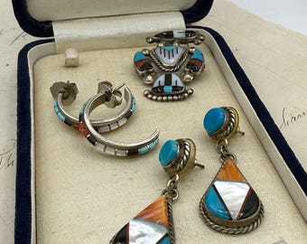 May include: A collection of silver jewelry including a brooch, a pair of hoop earrings, and a pair of teardrop dangle earrings. Each piece features inlaid segments of turquoise, white shell, orange stone, and black onyx in metal settings.