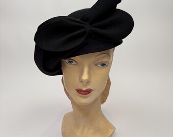 Puede incluir: Un sombrero de fieltro negro que presenta un lazo grande y estructurado en la parte delantera. El sombrero tiene una copa redondeada y un ala ligeramente asimétrica que se curva hacia arriba en un lado, con un diseño liso.