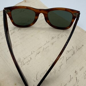 May include: Classic tortoiseshell sunglasses with green lenses. The arms are dark brown. The glasses are displayed on a piece of aged paper with handwritten script.