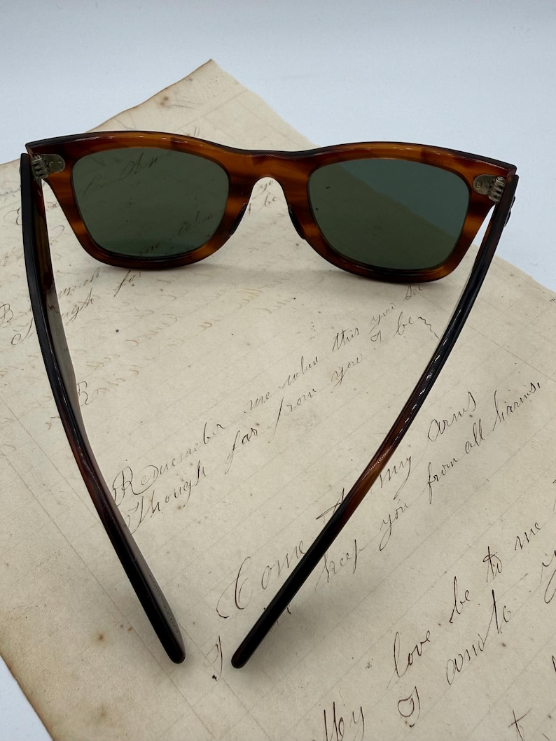 May include: Classic tortoiseshell sunglasses with green lenses. The arms are dark brown. The glasses are displayed on a piece of aged paper with handwritten script.