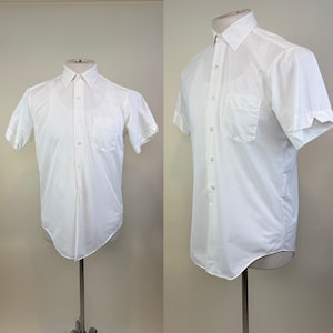 May include: A crisp white short-sleeve button-down shirt featuring a classic point collar, a single chest pocket on the left side, and a full-length front button placket. The shirt is displayed on a mannequin, showing a clean, structured design.