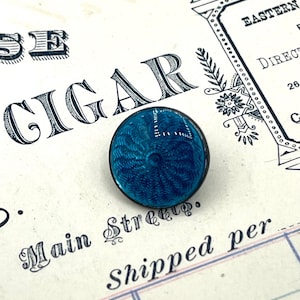 May include: A circular, turquoise enamel button featuring a detailed, swirling design. The button has a silver-coloured metal rim and is set against a vintage paper background with the word "CIGAR" printed in large letters.