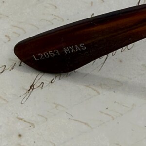 May include: Close-up of the arm of a pair of spectacles. The arm has a dark brown tortoiseshell pattern. The text "L2053 NXAS" is printed in white on the arm.