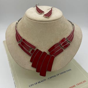 May include: A red and silver necklace and earring set. The necklace features a series of rectangular red enamel segments. The earrings are a matching design. The jewelry is displayed on a mannequin head, resting on a book with text.