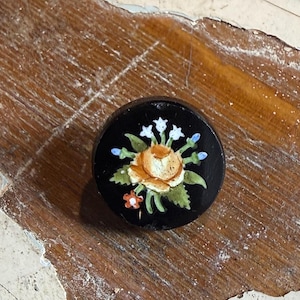 May include: A round, black decorative knob with a hand-painted floral design. The floral design features a yellow rose, white and blue accent flowers, and green leaves. The knob is likely made of wood or ceramic.
