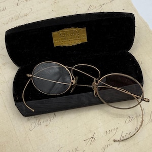Könnte beinhalten: Antike goldfarbene Brille mit runden Gläsern, in einem offenen schwarzen Samtetui. Das Etui hat ein goldenes Etikett mit Text. Die Brille liegt auf einem Blatt gealtertem Papier mit handschriftlichem Text.