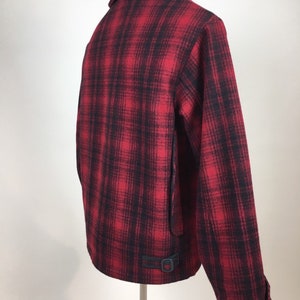 May include: A red and black plaid jacket with a collar and a buckle detail at the waist. The jacket is displayed on a stand, showcasing the back and side profile. The fabric appears to be a woven material.