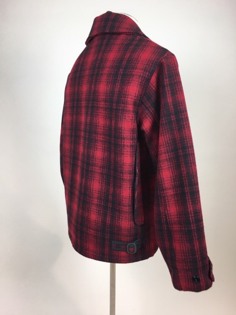 May include: A red and black plaid jacket with a collar and a buckle detail at the waist. The jacket is displayed on a stand, showcasing the back and side profile. The fabric appears to be a woven material.