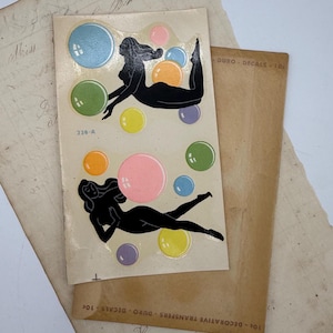 May include: Vintage decal sheet featuring two black silhouette figures with colorful bubbles. The figures are in a relaxed pose, surrounded by bubbles in shades of blue, green, yellow, orange, pink, and purple. The sheet is on aged paper.