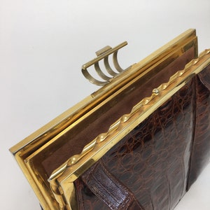 May include: A brown crocodile skin handbag with a gold metal frame and clasp. The handbag has a twisted gold metal trim around the edge of the frame.