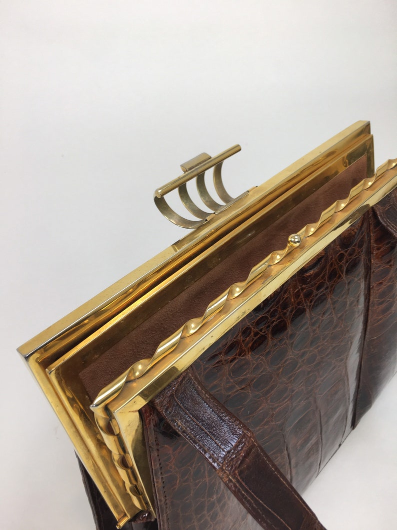 May include: A brown crocodile skin handbag with a gold metal frame and clasp. The handbag has a twisted gold metal trim around the edge of the frame.
