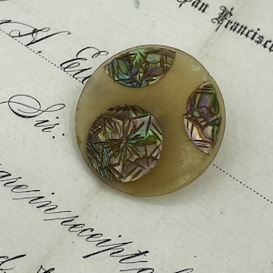 May include: A round, cream-colored button with three iridescent, circular designs. The designs are made of a material that resembles mother of pearl and have a geometric pattern.
