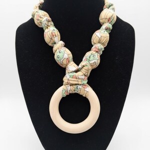 May include: A beige fabric necklace with a repeating cartoon character pattern, featuring a light wood ring pendant. The necklace is displayed on a black mannequin.