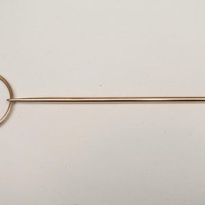 Op de afbeelding: Een koperkleurige haarspeld met een cirkelvormige handgreep en een lange, puntige pin. De handgreep heeft een decoratieve knoop. De haarspeld is ontworpen om haar vast te zetten en wordt waarschijnlijk gebruikt voor styling.