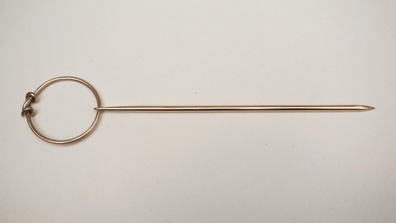 Op de afbeelding: Een koperkleurige haarspeld met een cirkelvormige handgreep en een lange, puntige pin. De handgreep heeft een decoratieve knoop. De haarspeld is ontworpen om haar vast te zetten en wordt waarschijnlijk gebruikt voor styling.