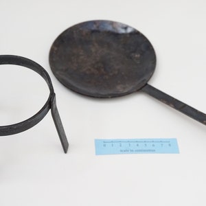 Puede incluir: Un soporte tr&iacute;pode de metal negro con una parte superior circular y una sart&eacute;n de metal negro con un mango largo. La sart&eacute;n est&aacute; ligeramente curvada y tiene un extremo redondeado. Una regla es visible en el fondo.