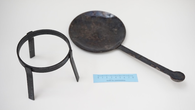 Puede incluir: Un soporte tr&iacute;pode de metal negro con una parte superior circular y una sart&eacute;n de metal negro con un mango largo. La sart&eacute;n est&aacute; ligeramente curvada y tiene un extremo redondeado. Una regla es visible en el fondo.