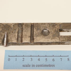 Può includere: Oggetto metallico rettangolare con una serie di elementi in rilievo e incassati. Presenta una sporgenza arrotondata, un'apertura rettangolare e una scala in centimetri. L'oggetto è lungo circa 8 cm.