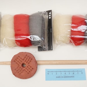 May include: Two clear plastic packages of carded wool in white, red, and dark grey, alongside a wooden drop spindle with a round, textured terracotta whorl. A blue ruler shows a scale in centimetres, measuring up to 8 centimetres in length.