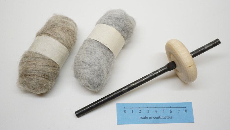 K&ouml;nnte beinhalten: Zwei Wollkn&auml;uel, eines hellbraun und eines grau, mit einem Holzspinnrad mit einer Metallspindel. Die Skala in Zentimetern ist in der rechten unteren Ecke des Bildes sichtbar.