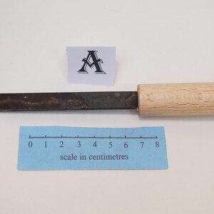 Puede incluir: Un cincel de metal con una hoja oscura y desgastada y un mango de madera clara. Una peque&ntilde;a tarjeta blanca con la letra A se sit&uacute;a sobre la herramienta. Una escala azul en cent&iacute;metros muestra que la herramienta mide 8 cent&iacute;metros.