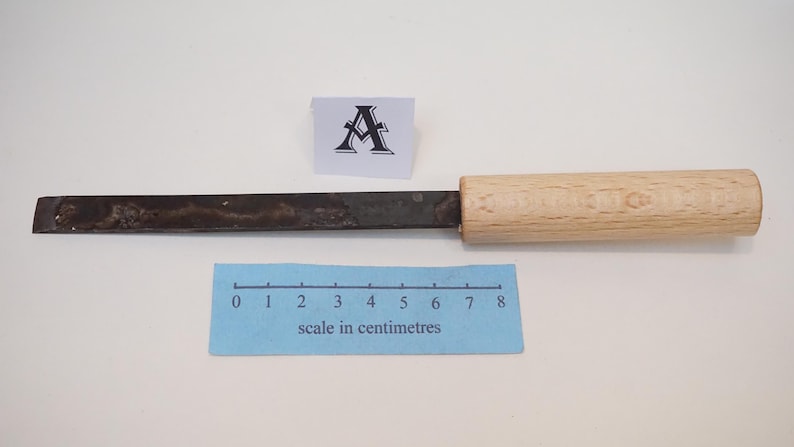 Puede incluir: Un cincel de metal con una hoja oscura y desgastada y un mango de madera clara. Una peque&ntilde;a tarjeta blanca con la letra A se sit&uacute;a sobre la herramienta. Una escala azul en cent&iacute;metros muestra que la herramienta mide 8 cent&iacute;metros.
