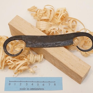 May include: A dark, hand-forged metal drawknife with curled handles resting on a rectangular block of light-coloured wood surrounded by wood shavings. A blue paper ruler shows the tool is approximately 8 centimetres in total length.
