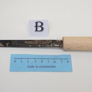 Puede incluir: Una gubia de metal para tallar madera con punta curva y mango de madera clara. Una cinta m&eacute;trica azul muestra que la herramienta mide aproximadamente 8 cent&iacute;metros de largo. Un peque&ntilde;o cuadrado blanco con la letra B se sit&uacute;a sobre la herramienta.