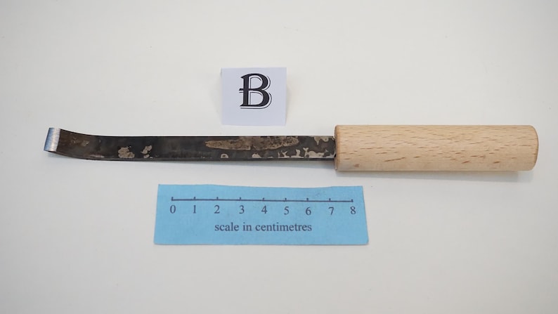 Puede incluir: Una gubia de metal para tallar madera con punta curva y mango de madera clara. Una cinta m&eacute;trica azul muestra que la herramienta mide aproximadamente 8 cent&iacute;metros de largo. Un peque&ntilde;o cuadrado blanco con la letra B se sit&uacute;a sobre la herramienta.