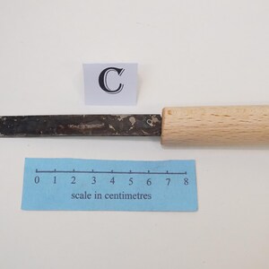 Puede incluir: Un cuchillo de manualidades con una hoja de metal oscuro y desgastado y un mango de madera clara. Un cuadrado blanco con la letra C se sit&uacute;a sobre la hoja. Una escala azul en cent&iacute;metros muestra que la herramienta mide 8 cent&iacute;metros.