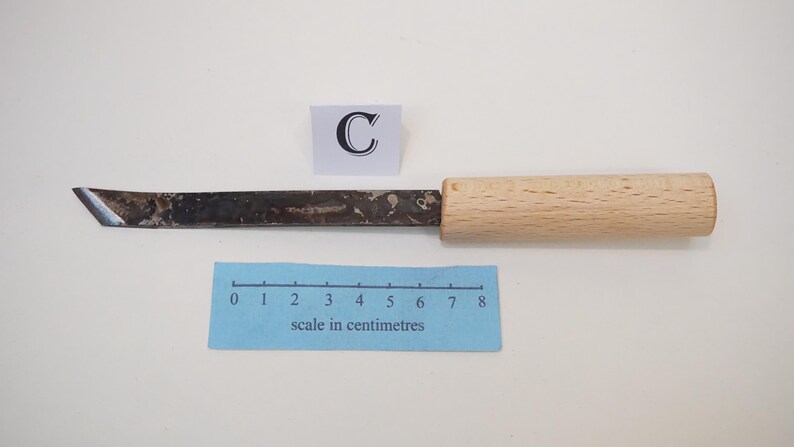 Puede incluir: Un cuchillo de manualidades con una hoja de metal oscuro y desgastado y un mango de madera clara. Un cuadrado blanco con la letra C se sit&uacute;a sobre la hoja. Una escala azul en cent&iacute;metros muestra que la herramienta mide 8 cent&iacute;metros.