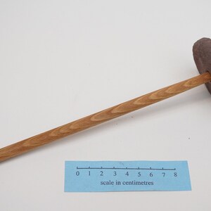 May include: A wooden spinning wheel with a brown clay whorl. The wheel is approximately 18 centimeters long.  The scale in centimeters is visible in the image.
