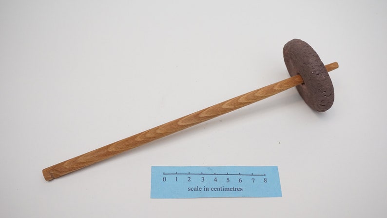 May include: A wooden spinning wheel with a brown clay whorl. The wheel is approximately 18 centimeters long.  The scale in centimeters is visible in the image.