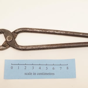 May include: A pair of dark grey metal blacksmith tongs with a pivot point and elongated handles. A blue paper scale is positioned below the tool, showing a measurement of 8 centimetres in length for the scale's marked range of units.