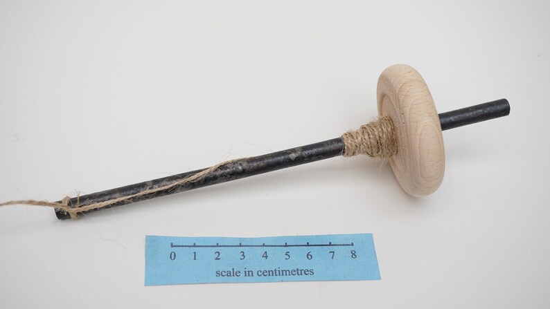 K&ouml;nnte beinhalten: Ein Holzspinnrad mit einer Metallspindel und einer L&auml;nge von Garn, das um sie gewickelt ist. Die Spindel ist etwa 20 cm lang.