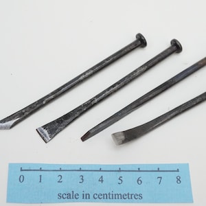 Peut inclure: Quatre outils métalliques avec des manches gris foncé et des têtes noires. Les outils ont des extrémités de formes différentes, dont un ciseau et un pied-de-biche. Une règle bleue avec le texte "scale in centimetres" est en bas.