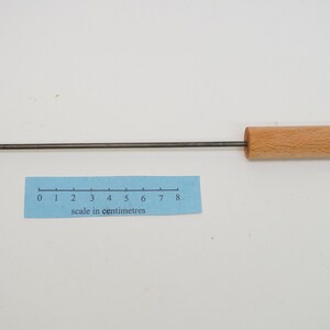 Pu&ograve; includere: Uno strumento di metallo lungo e sottile con un manico di legno. La parte metallica &egrave; piegata a forma di gancio a un'estremit&agrave; e ha un piccolo anello all'altra estremit&agrave;. Il manico di legno &egrave; lungo circa 8 centimetri. Lo strumento viene misurato con un righello che mostra i centimetri.