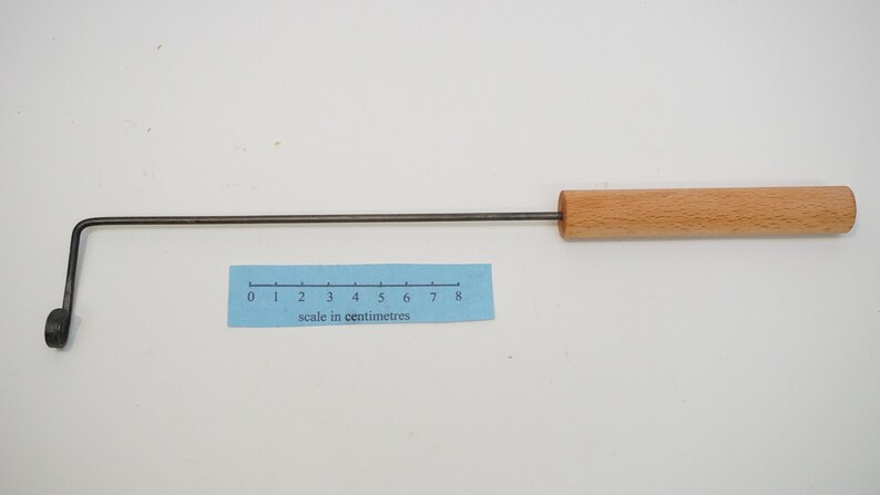 Pu&ograve; includere: Uno strumento di metallo lungo e sottile con un manico di legno. La parte metallica &egrave; piegata a forma di gancio a un'estremit&agrave; e ha un piccolo anello all'altra estremit&agrave;. Il manico di legno &egrave; lungo circa 8 centimetri. Lo strumento viene misurato con un righello che mostra i centimetri.