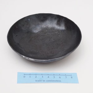 Puede incluir: Un cuenco de metal negro con un borde ligeramente irregular. El cuenco tiene un di&aacute;metro de aproximadamente 8 cent&iacute;metros.