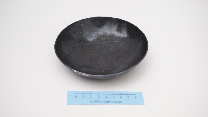 Puede incluir: Un cuenco de metal negro con un borde ligeramente irregular. El cuenco tiene un di&aacute;metro de aproximadamente 8 cent&iacute;metros.